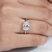 Oval-cut diamond engagement ring in a classic solitaire setting with a thin white metal band, displayed on a person's finger. The elongated diamond is secured by four prongs and showcases a clean, minimalist design.