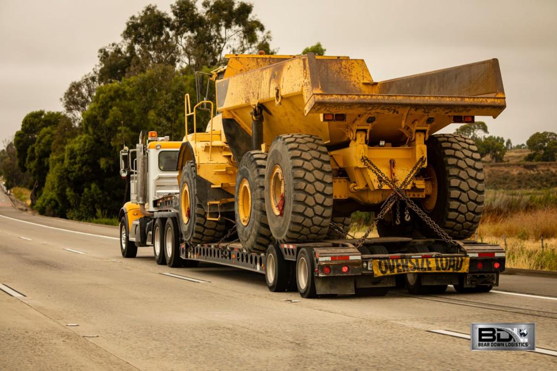 Bear Down Logistics transporting a large construction vehicle on a flatbed truck labeled 'Oversize Load' on a highway.